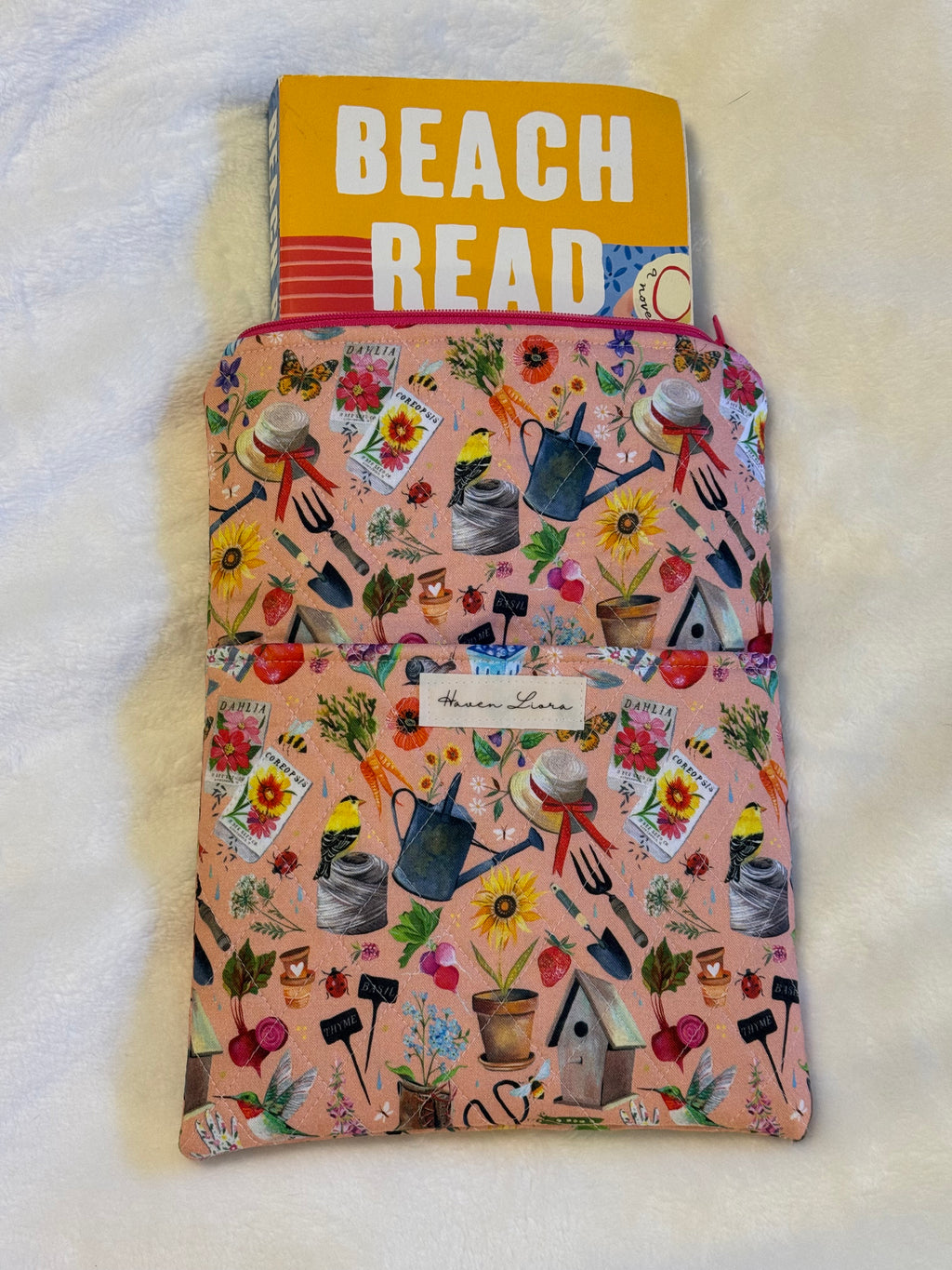 In the Garden Paperback Sleeve exterior in blush pink garden print with watering cans, seed packets, birdhouses and sunflowers